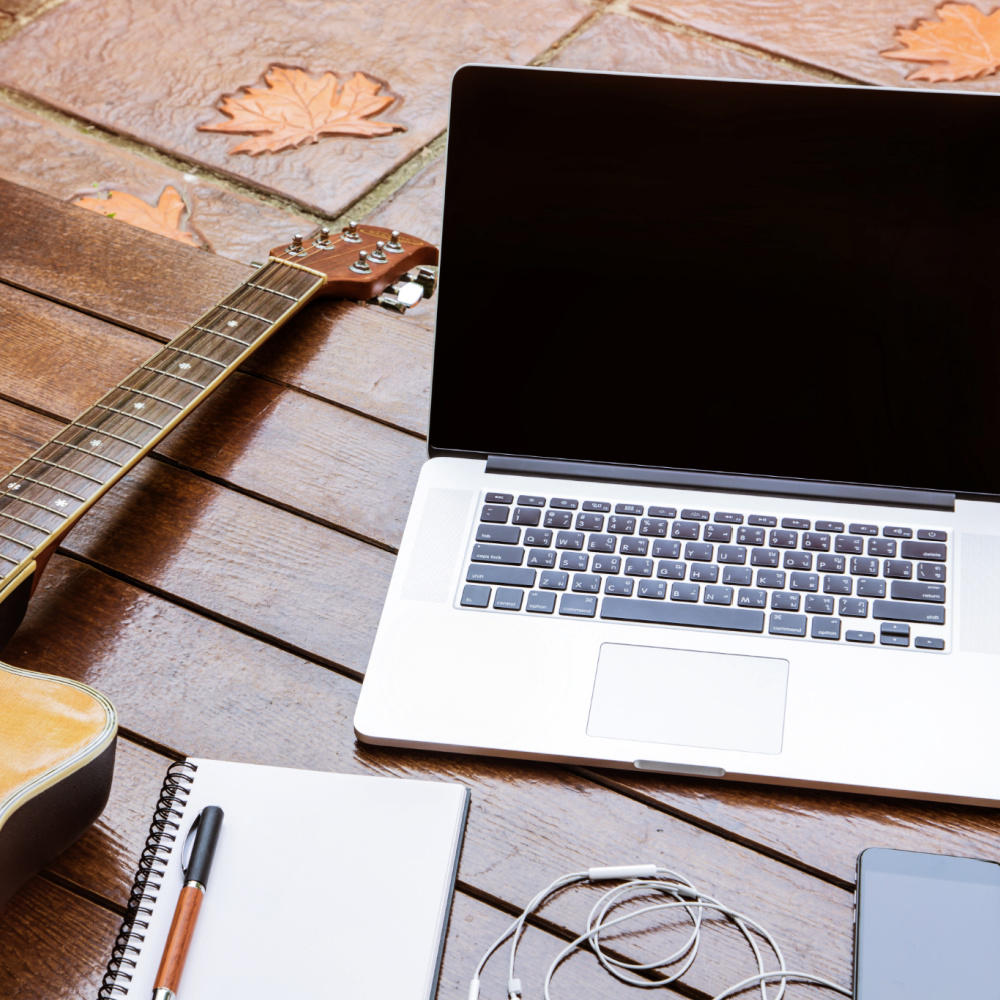 A laptop computer and acoustic guitar laying next to each other on a wood table, along with a notebook, pen, earbuds, and smart phone.