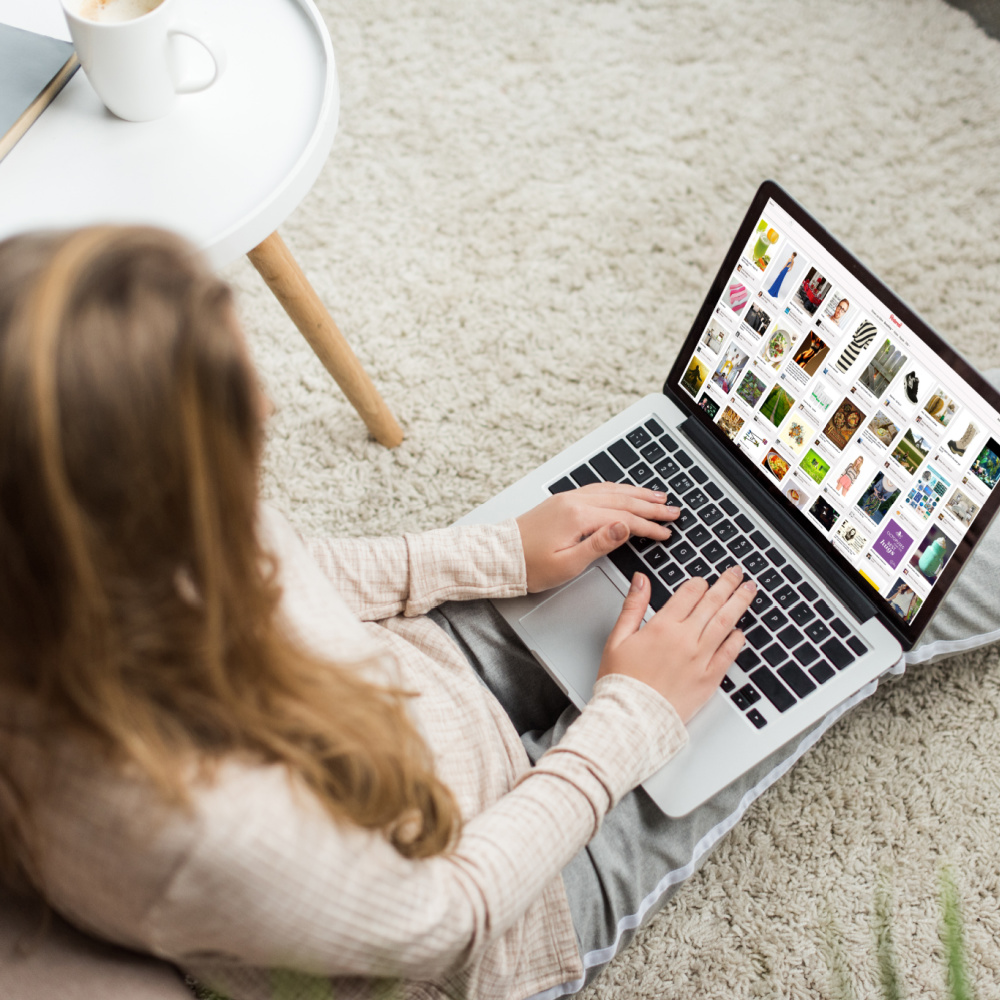 A woman with light brown hair sitting on the floor with a laptop computer, searching the home feed of Pinterest.