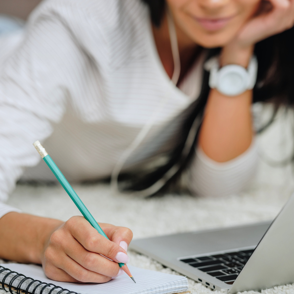 A young woman works on a laptop computer and writes with a pencil in a blank grid notebook.