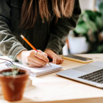 A young woman stands at a desk, writing in a notebook with a pencil. A laptop computer is open in front of her.