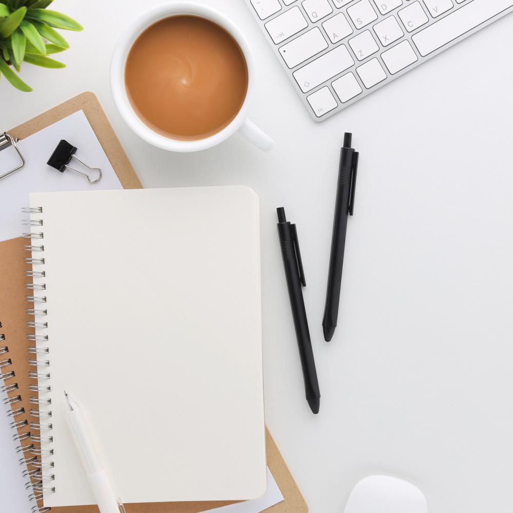 A blank notebook, a keyboard, succulent, coffee cup, pens, and a clipboard lay on top of a white desk