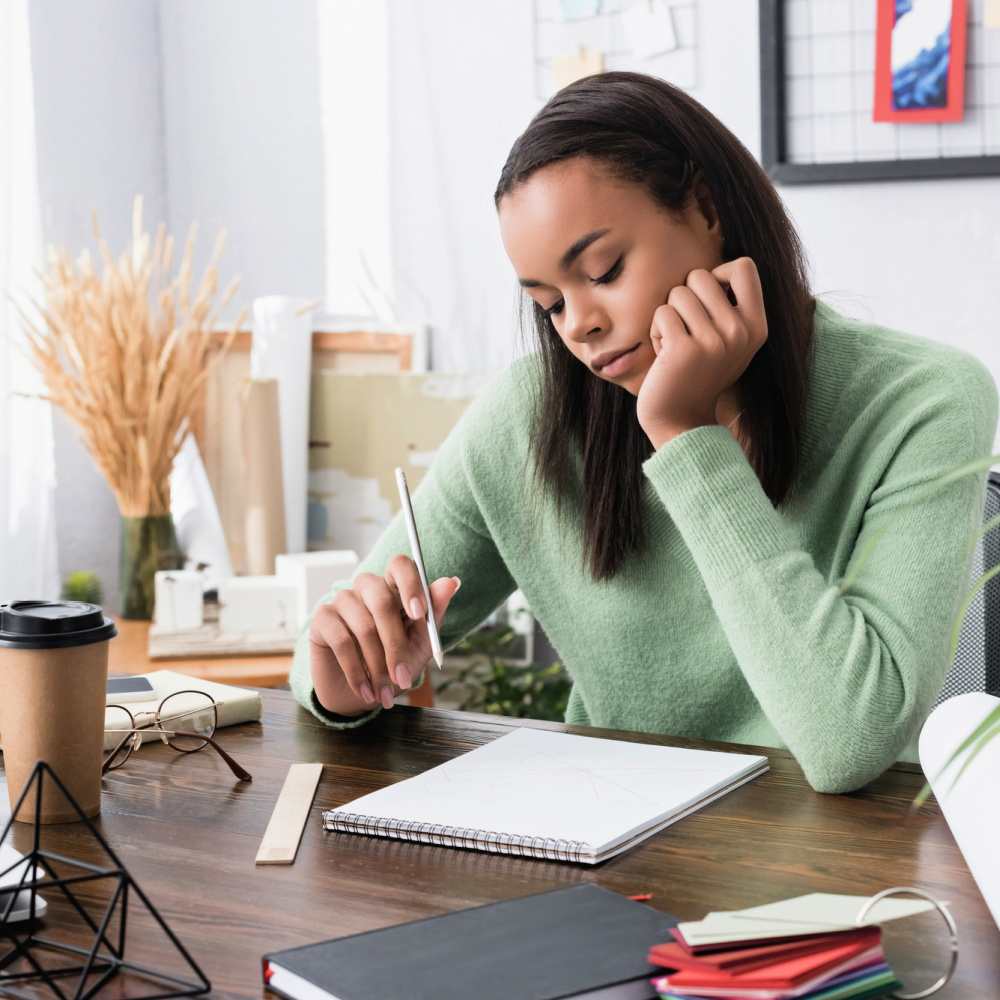 A young Black woman in a green sweater, sitting at a wood desk, looking at a blank notebook.