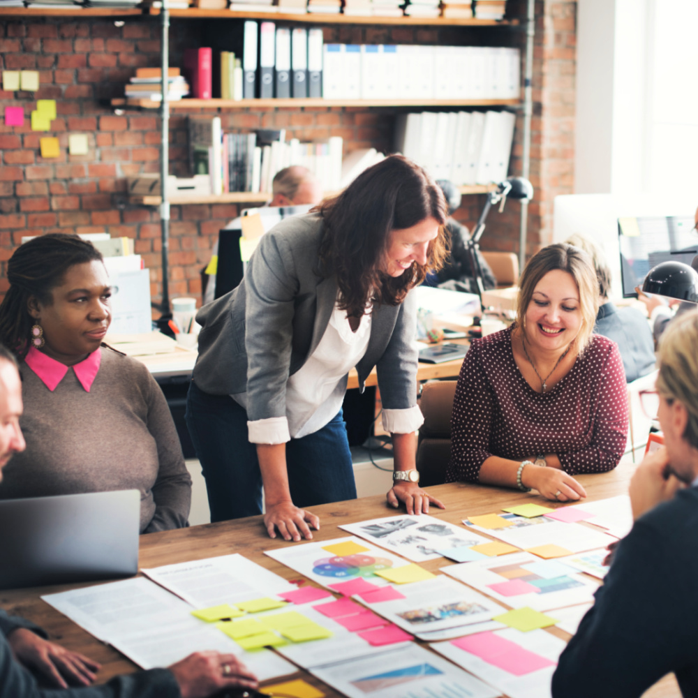 A group of people meeting and discussing at a large table, surrounded by papers, flyers, and sticky notes.