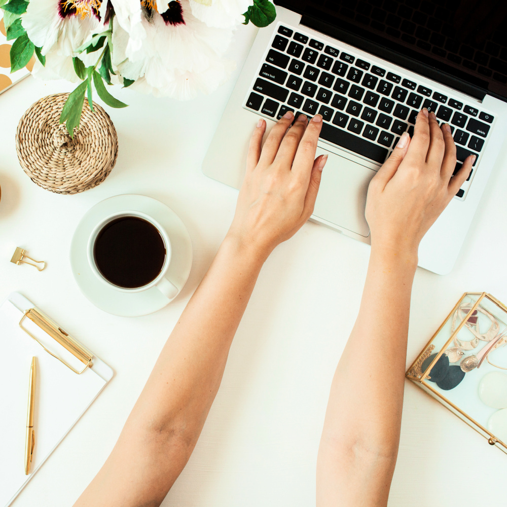 A woman's arms reach out to type on her laptop computer on a white desk. Accompanying the computer is a teacup with a beverage, a clipboard, pens, paper clips, and fresh flowers.